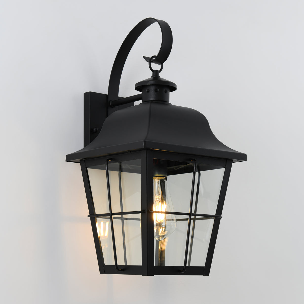 A black metal outdoor wall lantern with a curved top handle, clear glass panels, and two visible light bulbs inside, mounted on a plain white wall.