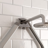Close-up of a modern stainless steel shower head and arm mounted on a white rectangular tiled bathroom wall. The shower fixture has the brand name artika engraved on it.