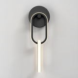 A modern wall sconce with a circular black base and an elongated oval light fixture, emitting a soft white glow, mounted on a plain light gray wall.