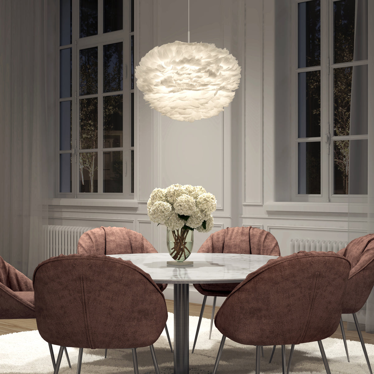 A modern dining room featuring a round marble table, six mauve upholstered chairs, a vase of white flowers, large windows, and the Artika Plume LED pendant light hanging above.