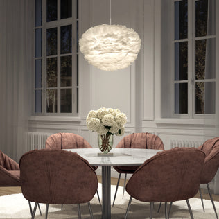 A modern dining room featuring a round marble table, six mauve upholstered chairs, a vase of white flowers, large windows, and the Artika Plume LED pendant light hanging above.