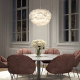 A modern dining room with a round marble table, pink upholstered chairs, a vase of white flowers, large windows, and a white, feathery pendant light hanging above the table.