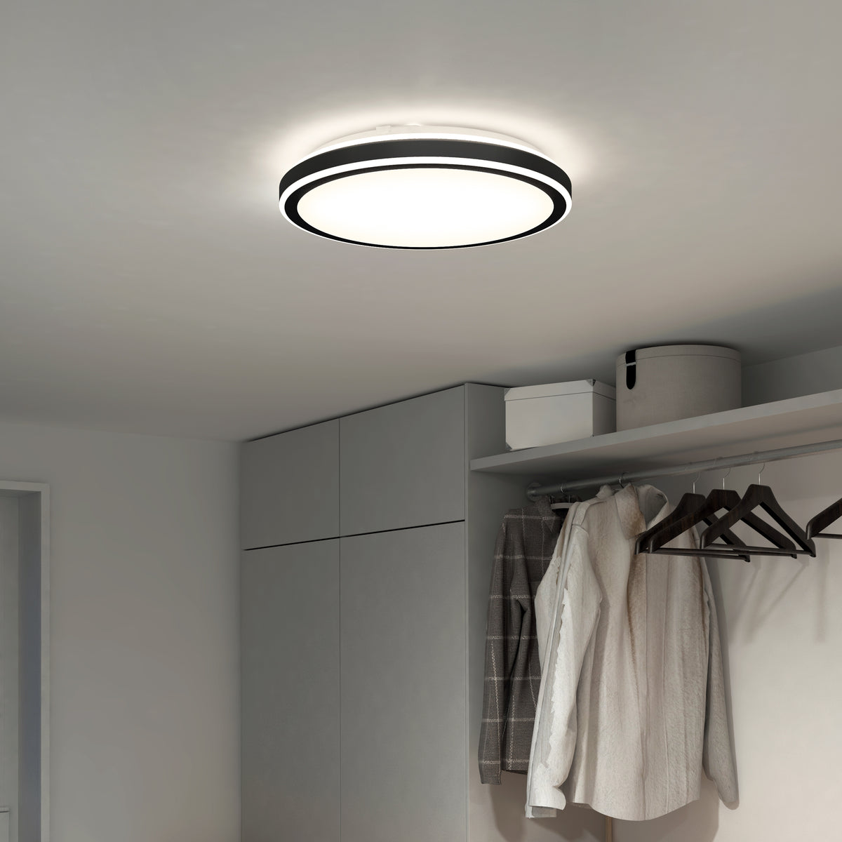 A modern round ceiling light fixture illuminates a minimalist closet with gray cabinets, hanging clothes, and storage boxes on an upper shelf. The space is bright and neatly organized.