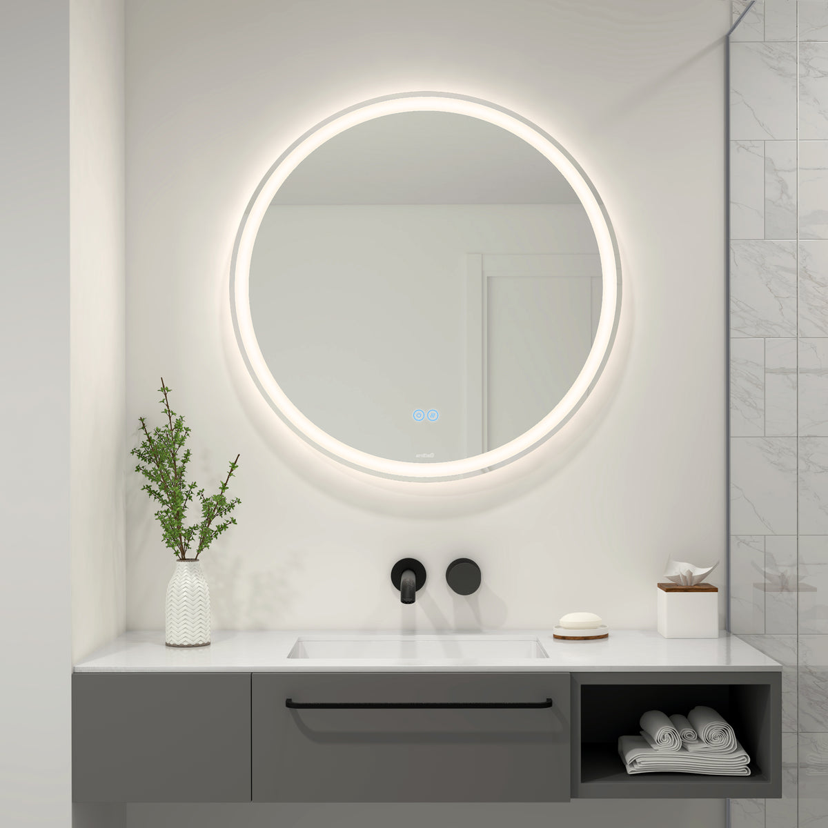 A modern bathroom features a round, illuminated mirror above a white sink with a black faucet. A vase with greenery, a soap dish, and a tissue box sit on the countertop, with marble tiles on the right wall.