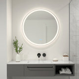 A modern bathroom features a round, illuminated mirror above a white sink with a black faucet. A vase with greenery, a soap dish, and a tissue box sit on the countertop, with marble tiles on the right wall.