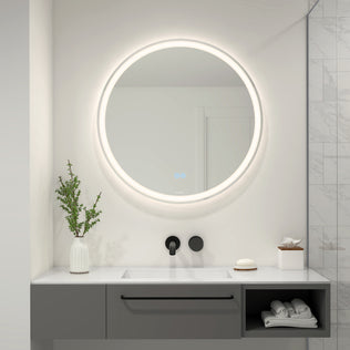 A modern bathroom features the Ayla 28 in. LED mirror 3 CCT with defogger by Artika above a sleek gray vanity. On the counter are a vase with greenery, a soap dish, and tissues. The wall is partly tiled with marble.