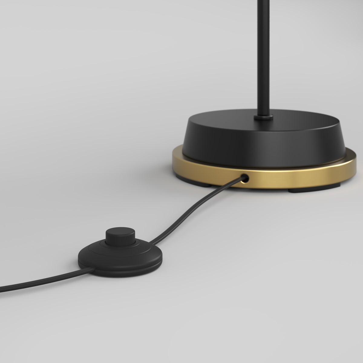 A close-up of a black and gold lamp base with a black cord attached, featuring a round inline foot switch on a light gray background.
