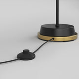 A close-up of a black and gold lamp base with a black cord attached, featuring a round inline foot switch on a light gray background.