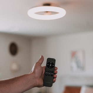 A hand holds the Artika Darren 23 in. LED fandelier ceiling fan remote, pointing at the modern fan with light.