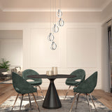A modern dining room with a round black table, four green velvet chairs, geometric pendant lights, wood flooring, and white paneled walls. Sunlight enters through a large window with sheer curtains.