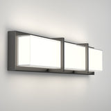 A modern wall-mounted light fixture with three rectangular frosted glass shades and a sleek metallic frame, emitting soft white light against a plain light-colored wall.