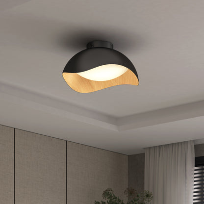 The Artika Adara 15 in. LED flush mount light features a black and wood-toned curved shade, casting a soft glow as it mounts on a white ceiling in a modern room with light walls and sheer curtains.