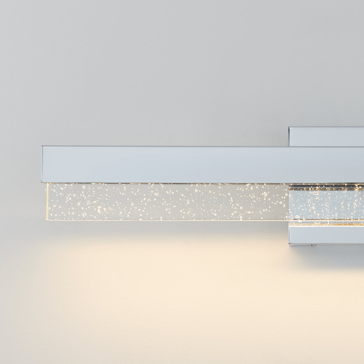 A modern wall-mounted light fixture with a sleek chrome finish and a rectangular glass bar embedded with small bubbles, emitting a soft white glow against a light gray wall.