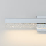A modern wall-mounted light fixture with a sleek chrome finish and a rectangular glass bar embedded with small bubbles, emitting a soft white glow against a light gray wall.