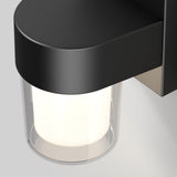 A close-up view of a modern wall-mounted light fixture with a black housing and a cylindrical frosted glass shade, emitting a soft white glow against a plain background.