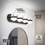 A modern bathroom light fixture with four globe-shaped LED lights mounted on a matte black bar above a round mirror. Text highlights Energy Efficient, Matte Black Finish, and a 3 Years Warranty.