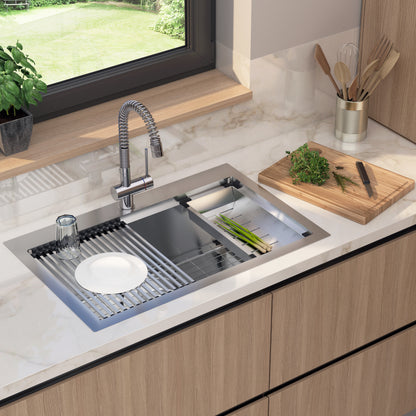 A modern kitchen sink with a flexible faucet, a drying rack holding a plate and glass, a cutting board with chopped herbs, and kitchen utensils in a holder. A window and potted plant are in the background.