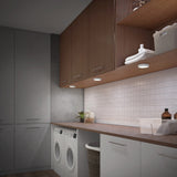 A modern laundry room with white cabinets, wood upper cabinets, a countertop, two front-loading washing machines, white tile backsplash, and baskets for storage. Under-cabinet lights illuminate the workspace.