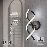 A modern wall light with two intertwined LED tubes in a matte black finish is mounted on a gray wall next to a mirror. Text highlights its energy efficiency, integrated LED, and 3-year warranty.