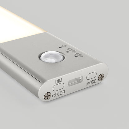 A close-up of the Artika Outline under cabinet LED light 3CCT shows its slim, rectangular design with a motion sensor and three labeled buttons—DIM, COLOR, and MODE—on a metallic surface. The LED is partially illuminated.