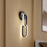 A modern wall sconce with two interlocking oval loops, one illuminated, mounted on a light wood wall next to a bed and abstract artwork.