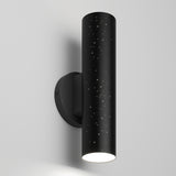 A modern black cylindrical wall sconce with tiny star-like perforations, mounted on a white wall, emits light from the bottom, creating a sleek and minimalist design.