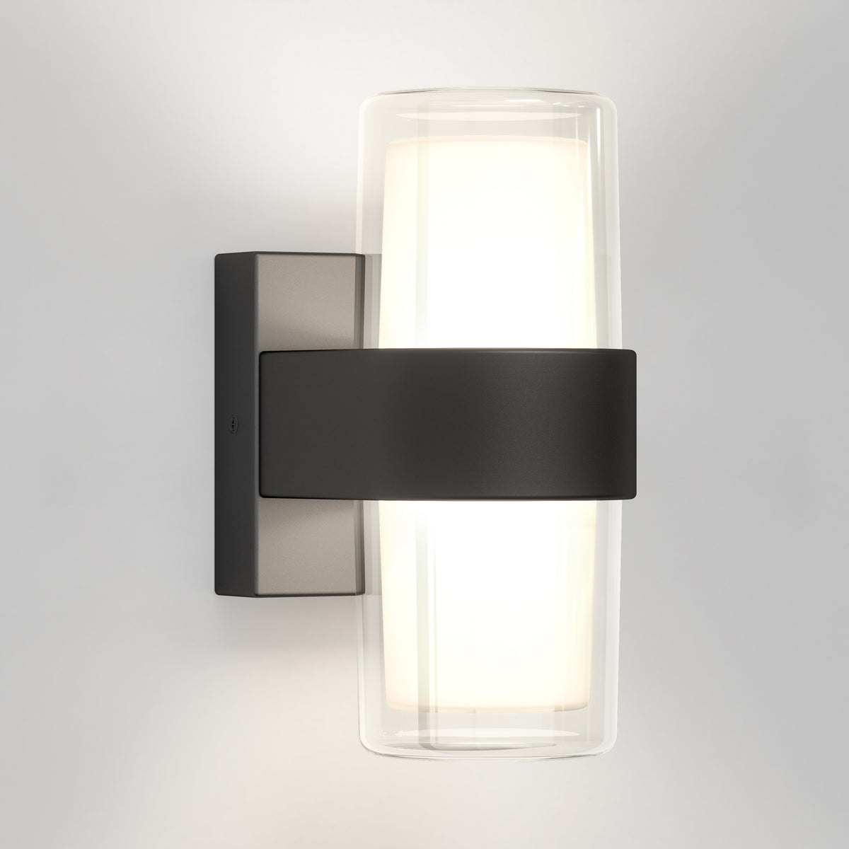 The Artika Colton modern LED indoor-outdoor wall light 5 CCT features a cylindrical frosted glass shade within clear glass, secured by a matte black metal band and rectangular base, and is shown mounted on a light-colored wall.