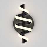A modern wall sconce with a black circular base and a vertical black rod, wrapped by a spiral LED light emitting a soft, warm glow against a plain light-gray background.