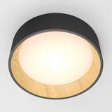 A modern ceiling light fixture with a black outer rim, wooden interior accent, and a glowing white circular light at the center, mounted on a white ceiling.