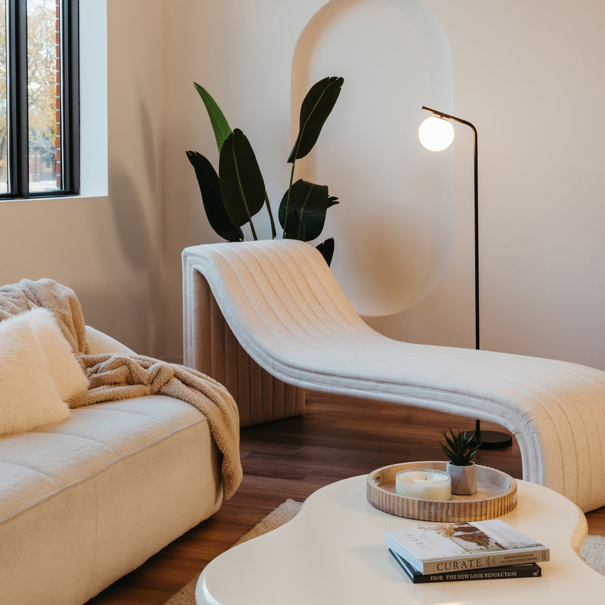 A modern living room with a sculptural cream chaise lounge, a white sofa with plush pillows and a beige throw, a white coffee table with books and decor, a floor lamp, indoor plants, and a large window.