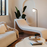 A modern living room with a sculptural cream chaise lounge, a white sofa with plush pillows and a beige throw, a white coffee table with books and decor, a floor lamp, indoor plants, and a large window.