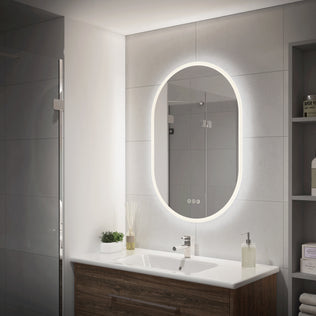 Modern bathroom with Artika Clara 30″x20″ oval LED mirror, white sink, wood cabinet, plant, soap dispenser, and toiletry shelves.