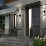 Modern house entrance with gray door, glass railing, and concrete steps. Two black and white wall lights flank the door, and small plants line the walkway in the foreground.