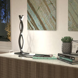 A modern table lamp with a twisted metal design sits on a marble surface, alongside books, a black beaded necklace, a small potted succulent, and abstract art hanging on the wall in the background.