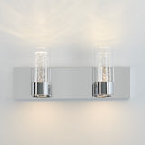A modern wall light fixture with two cylindrical glass shades, each containing tiny bubble-like details, mounted on a shiny rectangular chrome base, casting soft light on a white wall.