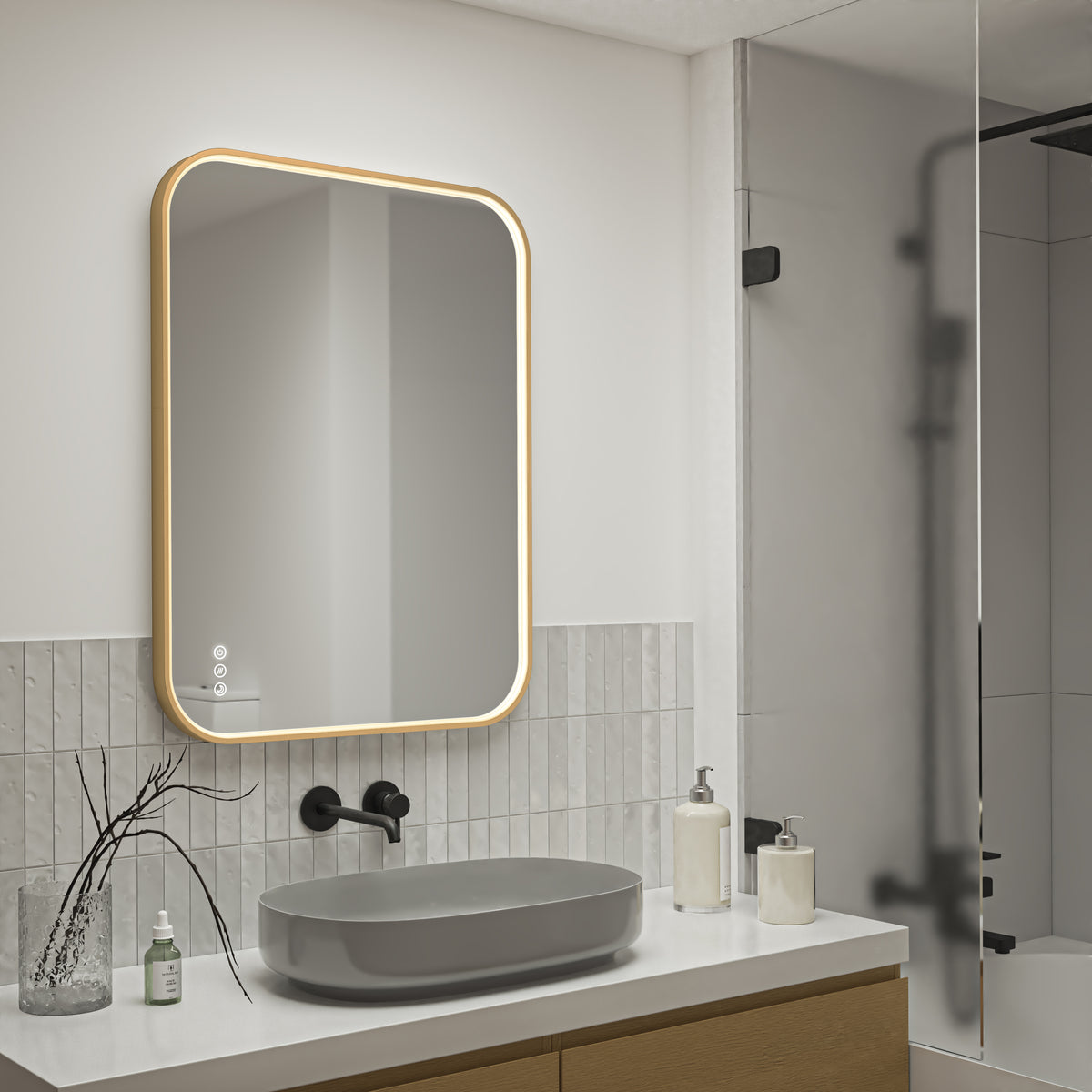 A modern bathroom showcases the Artika Royce Dimmable LED Mirror (3 CCT, horizontal/vertical mount) above a gray oval sink with a matte black faucet, soap dispensers, white tiles, wooden cabinetry, and a glass shower enclosure to the right.