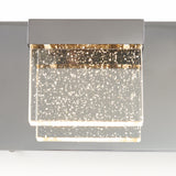 A modern square wall light fixture with a metallic base and a clear acrylic shade embedded with bubbles, illuminated and glowing warmly.