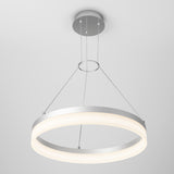 A modern circular LED pendant light with a silver frame, suspended from the ceiling by three thin cables, emits a soft white glow against a plain, light gray background.