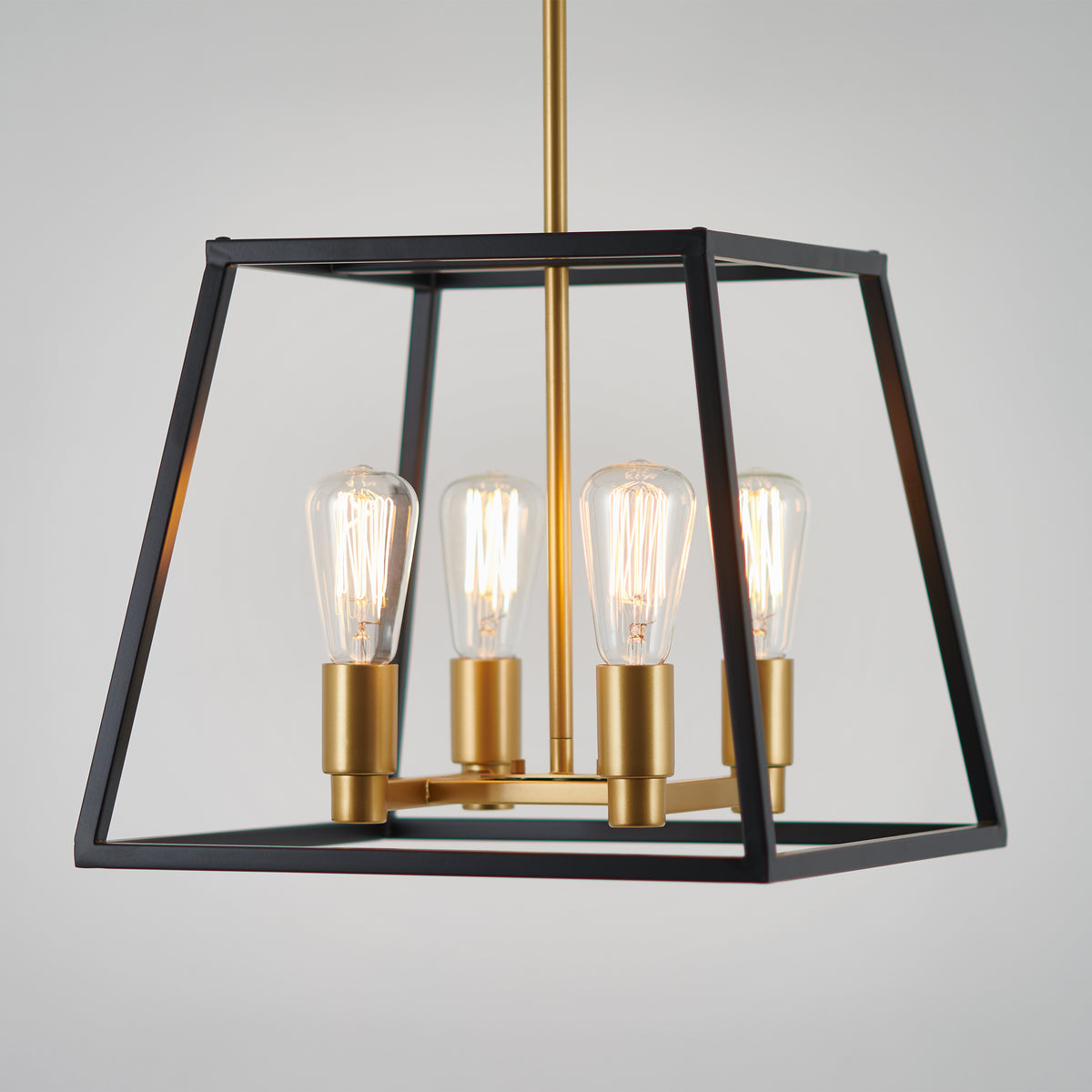 A modern pendant light fixture with a black open-frame geometric design, featuring four exposed Edison bulbs mounted on gold sockets, suspended from a gold rod against a plain background.