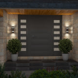 A modern front door with horizontal panels and frosted glass side accents, flanked by two wall-mounted lights and potted shrubs, set against a stone exterior and tiled entryway.
