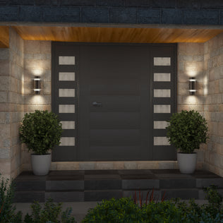 A modern front door with horizontal panels and rectangular glass inserts on each side, flanked by two potted shrubs and illuminated by a pair of Artika D4 indoor-outdoor wall lights mounted on a stone wall.
