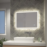 A modern bathroom with a rectangular, backlit mirror above a white vessel sink, wall-mounted faucet, soap dispenser, and tiled herringbone pattern wall. A window and small plant are on the left.