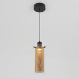 A modern pendant light with a cylindrical glass shade and an inner perforated metal tube, suspended from a black ceiling mount by a single black cord. The light is turned on, emitting a warm glow.