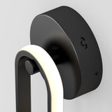 Close-up of a modern, black wall-mounted light fixture with a rounded rectangular design and a glowing white light strip, attached to a curved black base against a plain white wall.