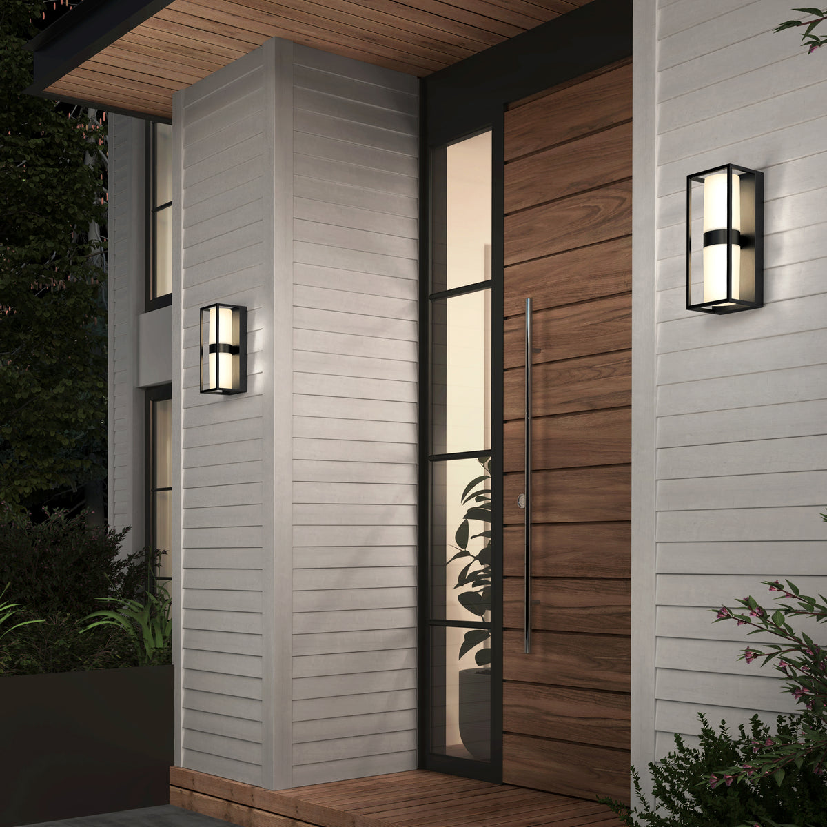Modern house entrance with vertical wooden door, large glass panel, and two rectangular wall-mounted light fixtures on light gray siding. Plants and greenery border the entryway.