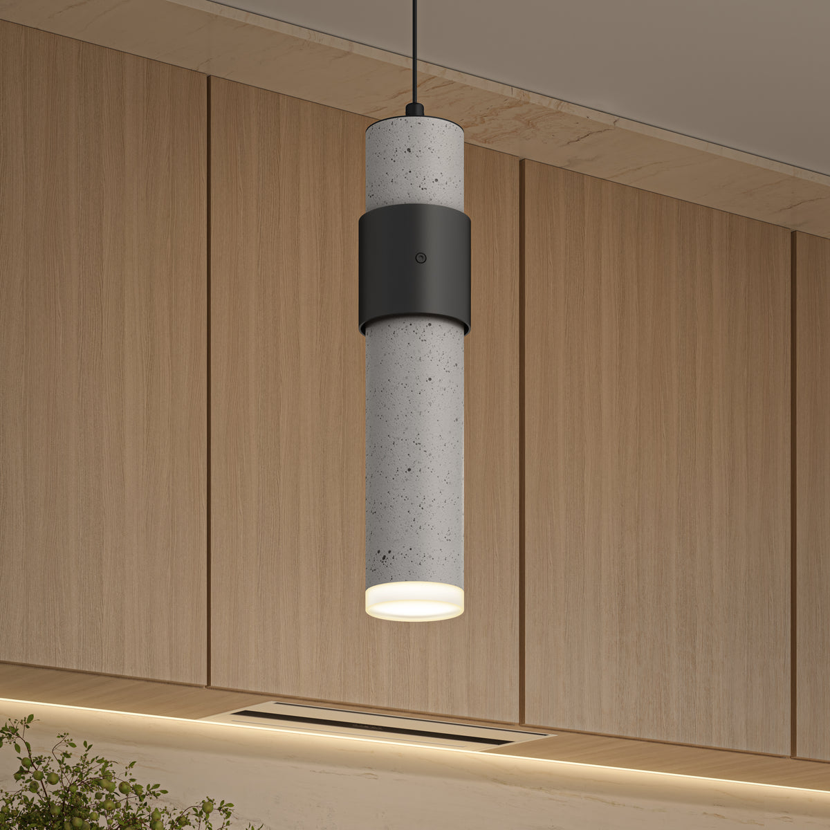 A modern cylindrical pendant light with a concrete texture and black accent hangs from the ceiling in front of light wood cabinets in a contemporary interior setting.
