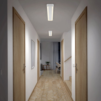A well-lit hallway with light wooden floors and four closed wooden doors, two per side. Artika's Aiden 48 in. LED modern flush mount ceiling light brightens the space. At the end, a room features a plant, chair, and large curtain.