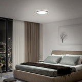 A modern bedroom with a large bed, gray bedding, floor-to-ceiling windows with city views, neutral curtains, and a round ceiling light. A framed tree artwork hangs above the bed.