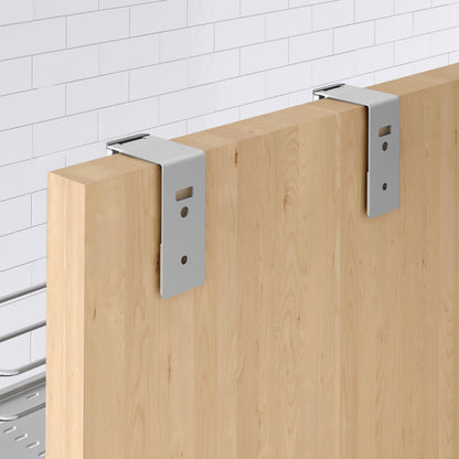 Two silver metal brackets are attached to the top edge of the Artika Camden Shower Caddy’s light wood panel, set against a white tiled wall. The brackets have screw holes and are designed for mounting or support.