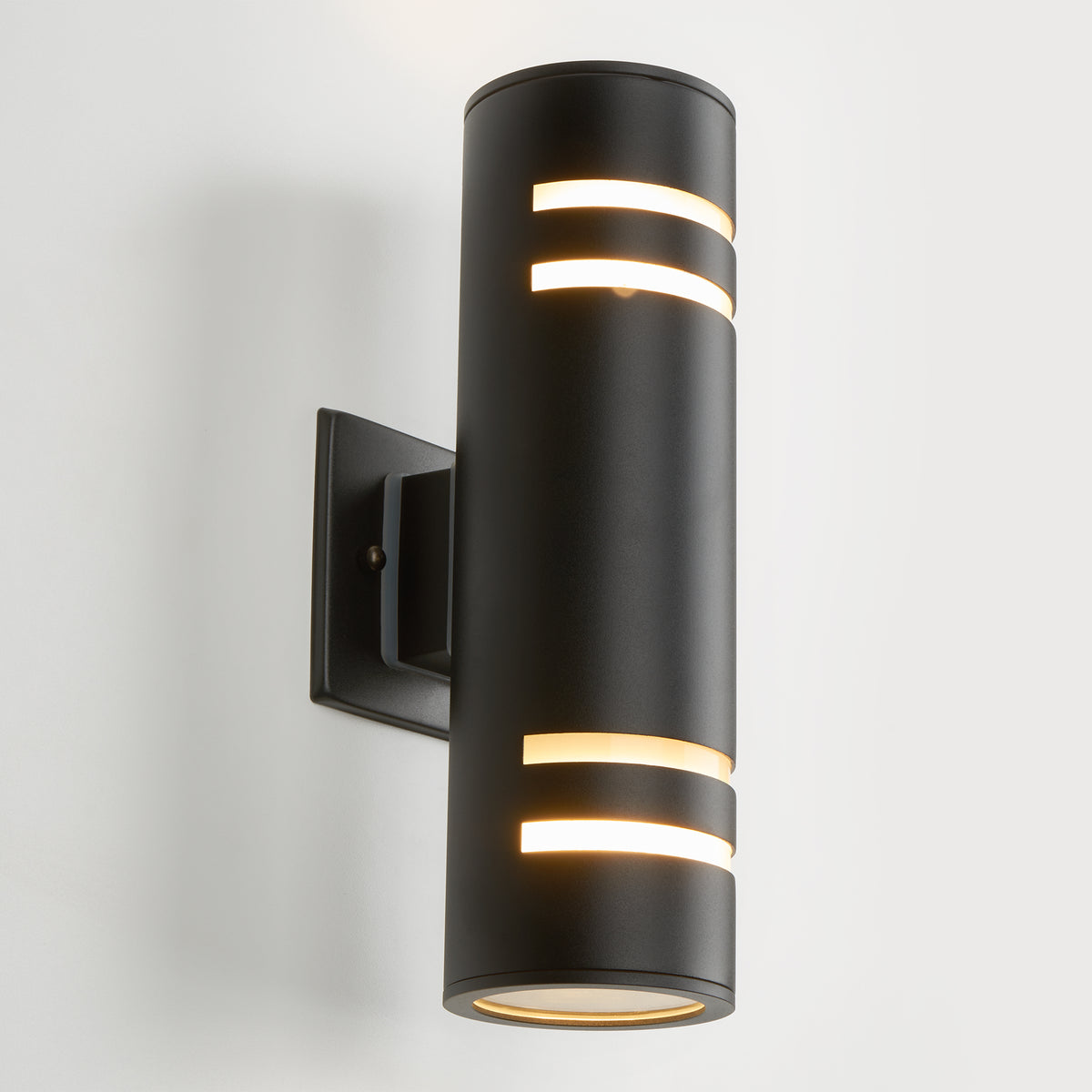 A modern black cylindrical wall sconce with horizontal light slits, mounted on a square base, emitting warm light from both the top, bottom, and side openings. The light is fixed to a white wall.